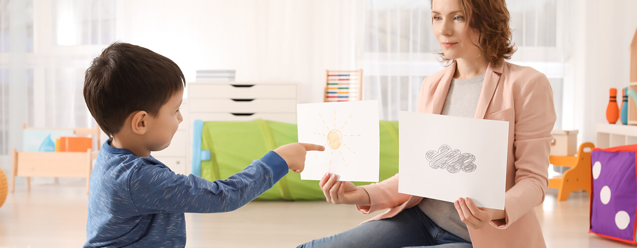 A therapist holds up a picture in each hand, while the child sitting across from them points to a picture.