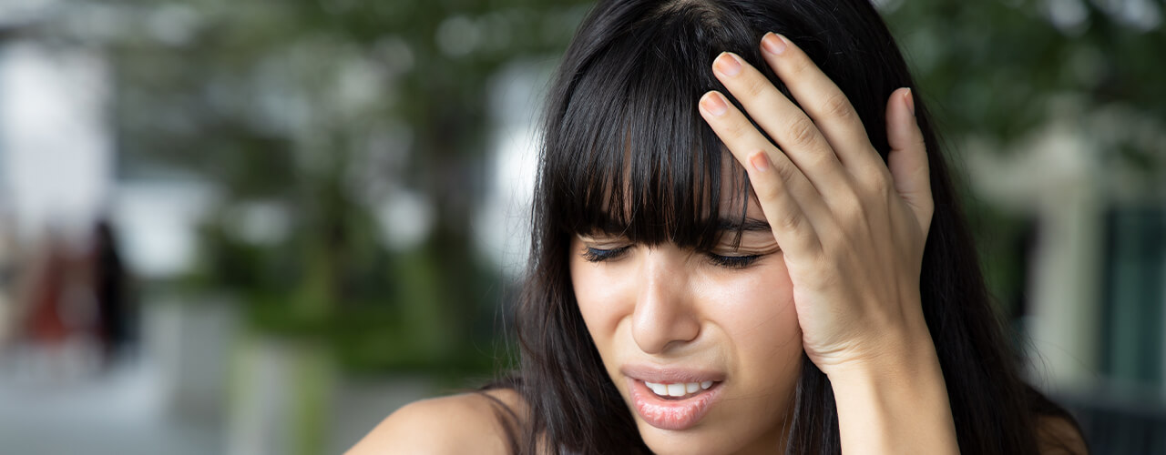 Girl with black hair has a hand on her head, with a face showing she's in pain.