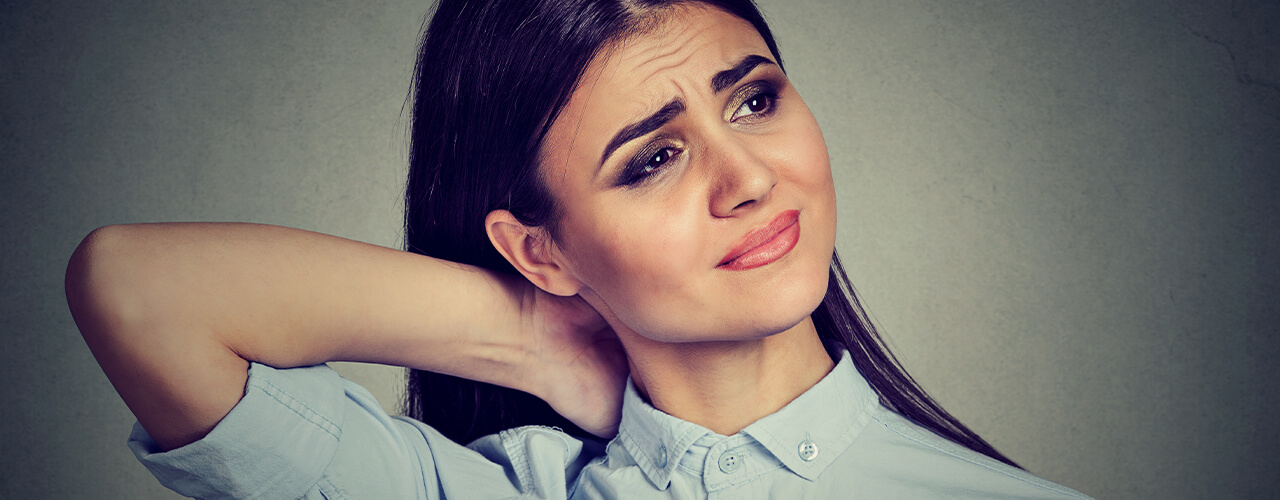 A woman with dark brunette hair reaches her right hand to grip the back of her neck.