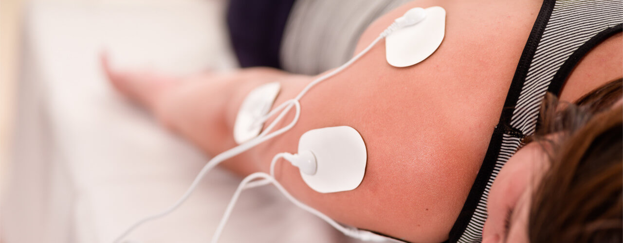 A woman lays on their stomach, while she receives iontophoresis treatment on her shoulder.