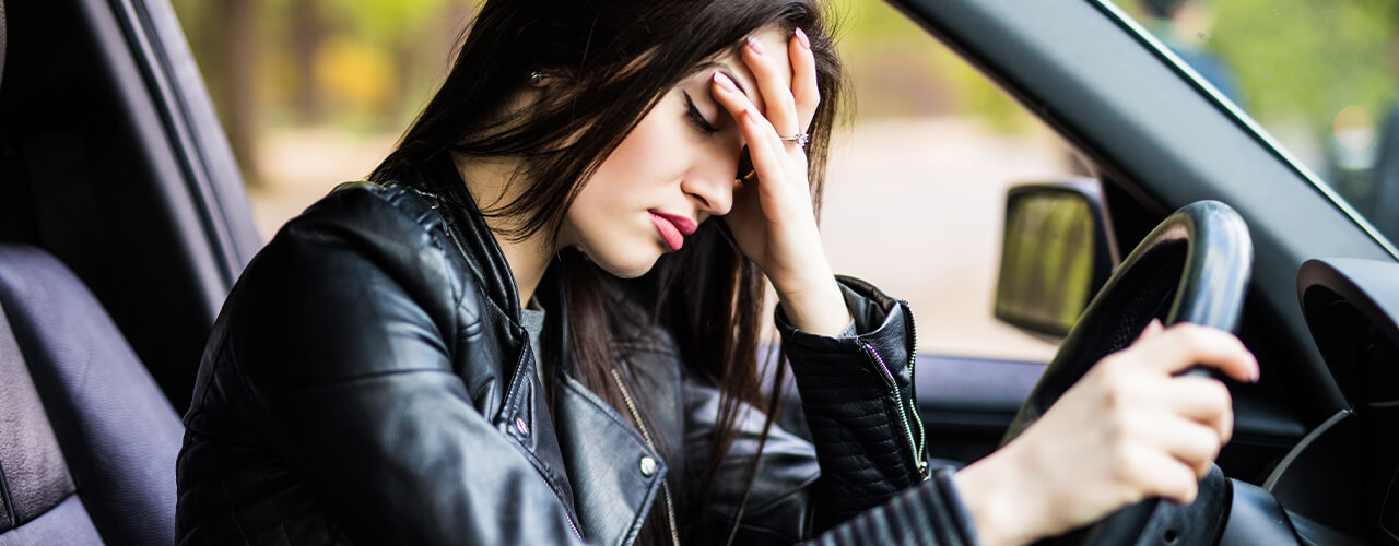 A woman with long brown hair sits in the drivers seat of a car with her right hand on the steering wheel and left hand on her forehead.