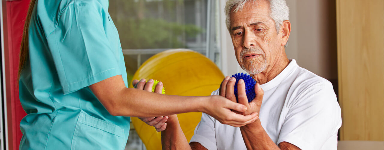 Older man sitting with a therapy ball in his hand, while a therapist assists him.