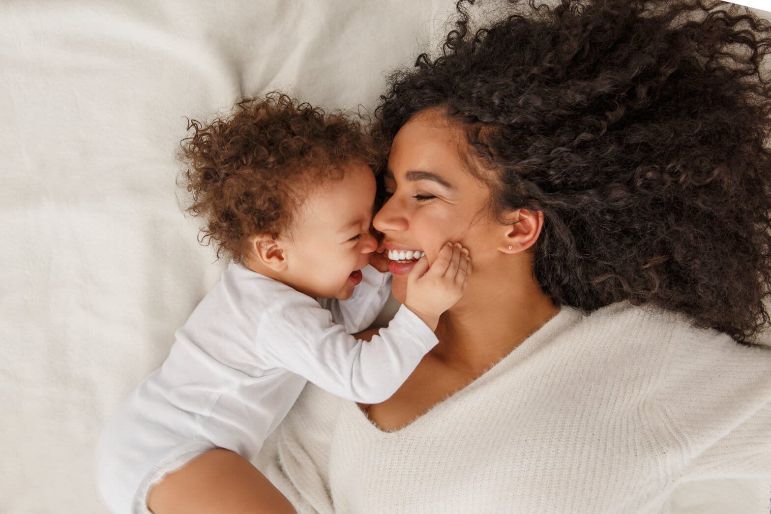A woman with dark curly hair lays in bed with her baby smiling as the baby has a hand on her cheek.